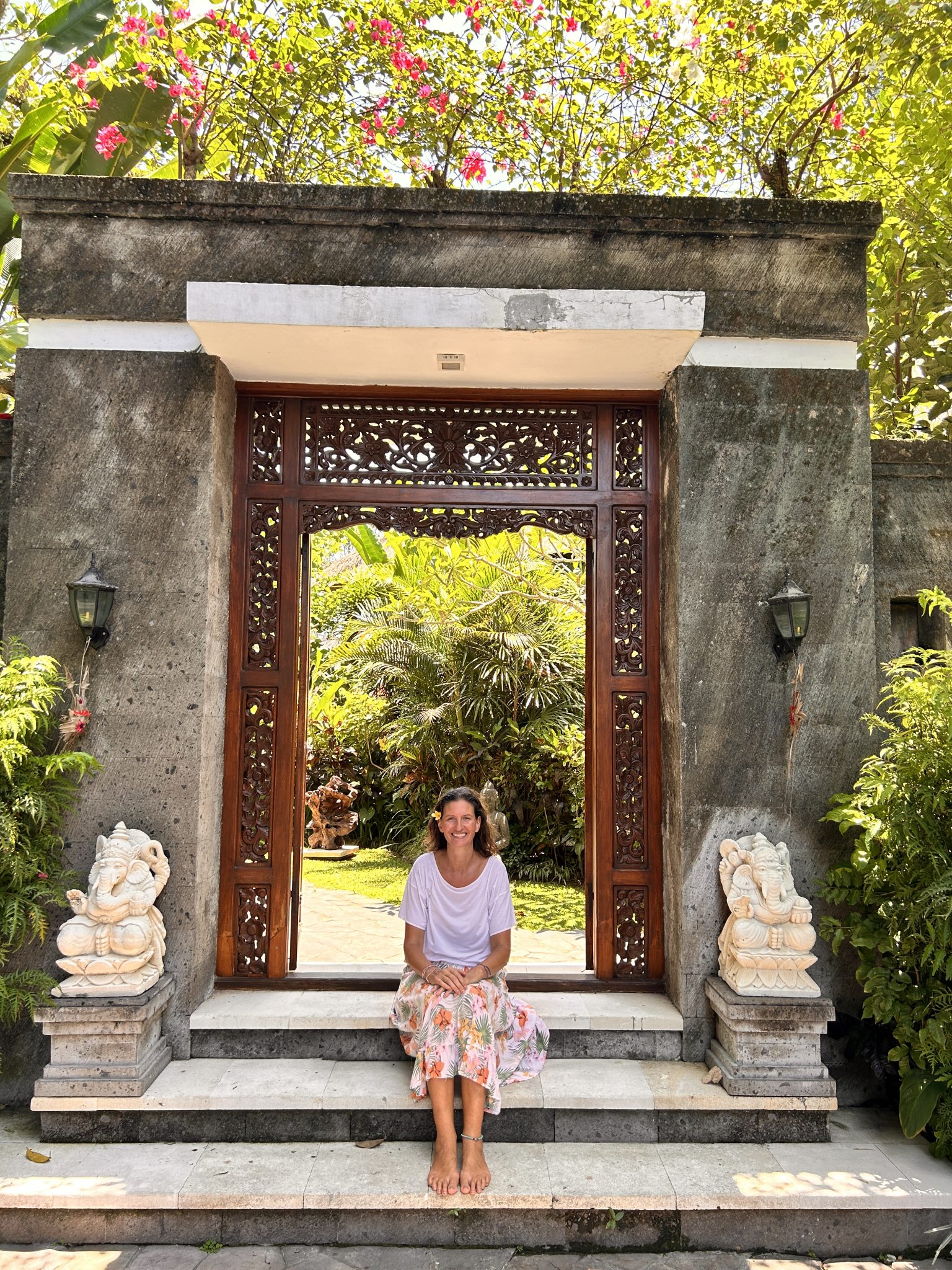 A woman stands on a path surrounded by lush green plants and trees, smiling at the camera. She is wearing a white shirt and a floral skirt, embracing the serene ambiance of her women’s retreat in Bali.